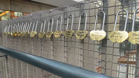 Tom Grundy/BBC Heart shaped golden padlocks on the bridge with names and ages.