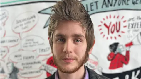 A photo of young dad James smiling for the camera. He has brunette hair that is in quite a long quiff at the front and has a small beard and moustache. It's a close up photo of his head and behind him is a mural on the wall with positive phrases on about being a dad.