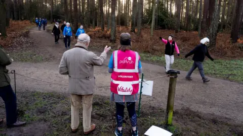 PA Media People running, jogging or walking through a muddy path. The King is standing facing them with his back to the camera.
People are waving at the King and he is waving back.
The King is wearing a brown zip up jacket. 