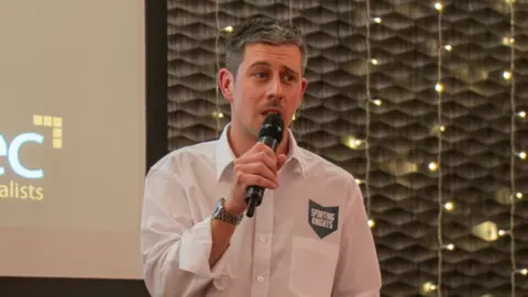 Gavin Caney Gavin Caney speaking into a microphone. He is wearing a white shirt and looking slightly off to the right of the camera.