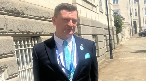 Thomas Mallon, stood outside by a large stone building. He is wearing a light blue tie and a suit.