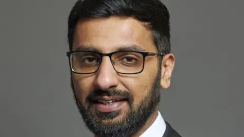 Official portrait of Adnan Hussain taken against a grey backdrop. He has black hair and a beard and is wearing glasses.