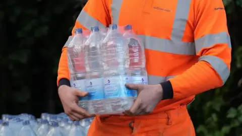 A man in an orange hi-vis jacket with a case of water bottles in his hands.