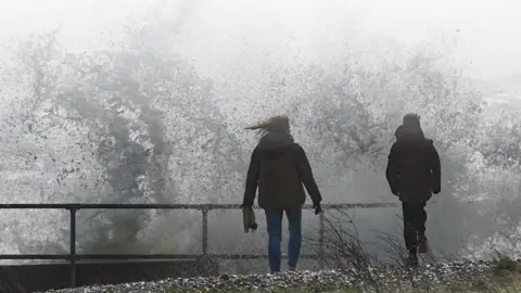 Coastal Partners Two people facing away from the camera, looking out to see. They are stood on top of a sea wall with waves crashing into them
