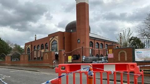 Manzil Way, with a mosque in the background and red crash barriers in the forefront of the picture, with police tape in front of it 