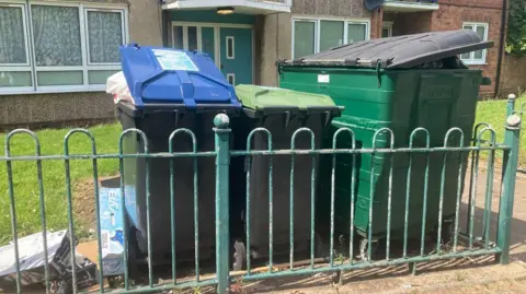 Three bins outside low-rise flats in, they are so full that their lids are unable to fully close, and there is cardboard left on the ground to the side of them