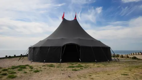 A large black tent which has been set up on a seafront.