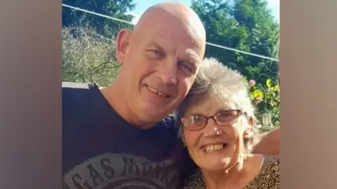 A man with a bald head, wearing a black t-shirt with a monkey on it, has his shoulder around an older woman with glasses and a printed shirt. Both are smiling in a garden on a sunny day.