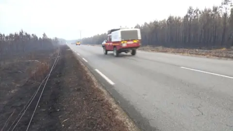 A firetruck travels down a road with burnt vegetation on either side