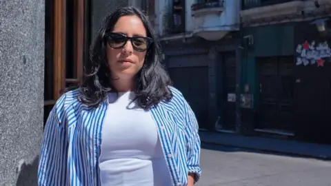 Submitter photo Alyssa wears a blue and white striped shirt over a white t-shirt and black sunglasses. She looks toward the camera on the corner of a street in Uruguay. 