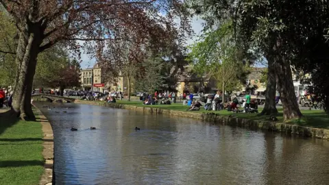 Getty Images People sitting in the sunshine along the waterside in Bourton-on-the-Water