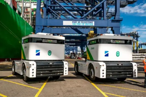 Port of Felixstowe Autonomous white trucks underneath blue metal towers at Felixstowe Port.