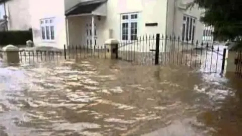 Local Democracy Reporting Service Flood water almost reaching the windows of a cream house and half-submerging the gates outside.