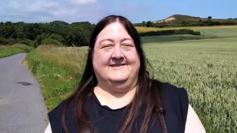 Margaret Crosby is standing in front of fields and there is a narrow road beside her. The Hastings Hill Round Barrow can be seen in the distance.