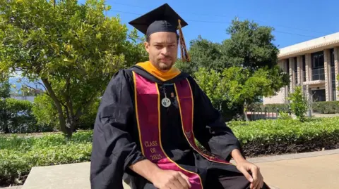 LinkedIn Cole Allen sitting on a wall outside in the sun in front of a grassy area with trees. He looks at the camera, and is wearing a graduation cap and gown. The red and yellow sash says class of 2025 on it. 