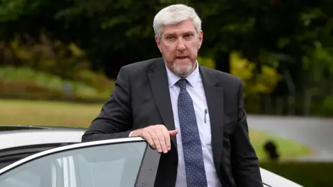 PA Media John O'Dowd (grey hair and beard) in black suit, blue tie and grey shirt