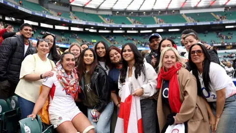 Getty Haineala Lutui with family and friends. She is wearing the England kit and a woman at the front has a flag. The group are smiling towards the camera. 