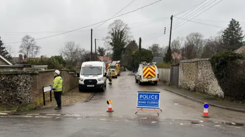 The entrance of a street with flint and brick walls on both sides. The road is closed and has a blue sign saying "Police, road closed" in white letters, between with two road cones. Beyond, parked, are emergency vehicles including a fire engine. A firefighter is on the left in the distance, and in the foreground is a police officer wearing a hi-vis jacket and a white hat.