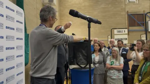 BBC The image shows the county's deputy returning officer drawing a piece of paper and holding it above a ballot box, on a stage surrounded by the candidates and other attendees. They are in a leisure centre sports hall with count signs and a basketball net visible.