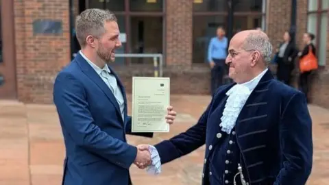 Keiran Sandham with Cumbria's High Sheriff outside Carlisle Crown Court. They are shaking hands and Mr Sandham is holding a letter. Mr Sandham has short grey hair and a beard and is wearing a blue suit. The High Sheriff is wearing a navy blue court dress.