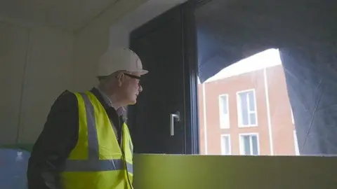 WMCA Mayor Richard Parker stands inside one of the new social homes under construction at Port Loop. He is looking out of a window at other buildings in the development. The room he is in is still being built. He wears a white hard hat and black rimmed glasses with a green high-vis vest over a dark suit jacket.