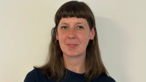 Photo of Sara against a neutral off-white background. She is looking directly at the camera. She has long brown hair and a fringe. Her hair is behind her ears and she wears hexagonal earrings. 