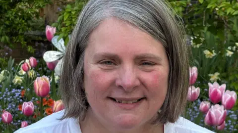 Vanessa Alderton A smiling Vanessa Alderton has a grey/brown bob hairstyle and a white top. Behind her are flowers in a garden.