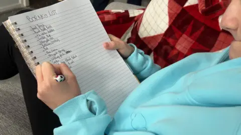 Milly is pictured writing a list of all the things she needs to take with her, including mosquito spray and a head torch.