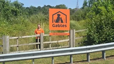 Claire Sparkes is pictured standing in front of an orange Gables sign. She is standing to the left of the sign, behind the fence which is overgrown with plants and trees.