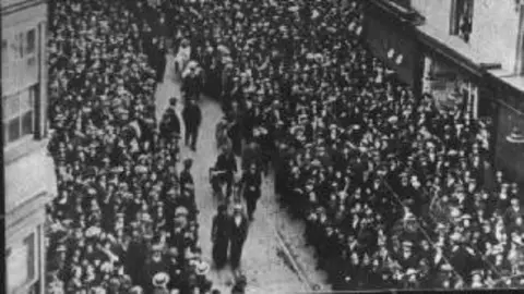 SCC Libraries Huge crowds gathered at the pagan hour of 6am to hear St Mary's choristers greet May Day from the top of the Bargate, from which this photograph was taken. A good view is given of William Lewis Parkhouse, jewellers, 10 Above Bar Street, the corner of Hanover Buildings and shops further along the east side of Above Bar Street. A feature of many crowd scenes of this date is the large number of people wearing hats.