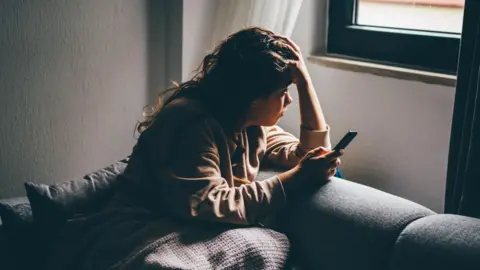 Woman appearing sad, holds her phone in one hand and her head in another. She is sitting on a sofa, wearing a blanket and looking out of the window