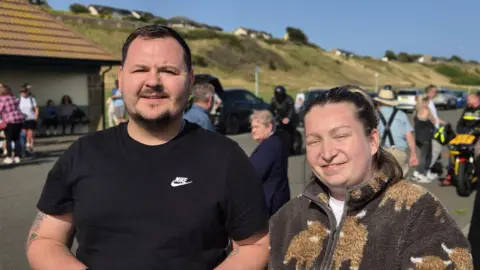 Kieran Esson and Megan Davies look at the camera with people behind them at a park