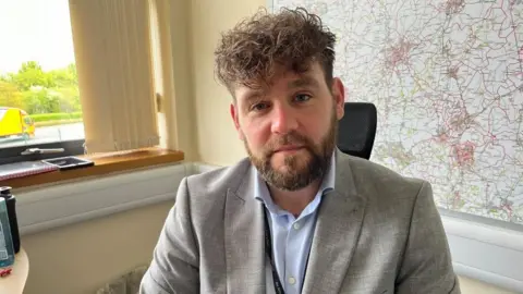 Ollie Conopo/BBC Gareth Chisholm - man with brown hair and beard sitting in front of a large map