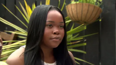 BBC Angel Mbondiya sits in a garden in front of a plant and hanging basket