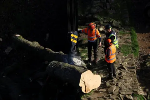 Reuters/Lee Smith Workers stand near a log, as sections of the Sycamore Gap tree, that was felled by a vandal last month, are removed from the area by crane in Northumberland