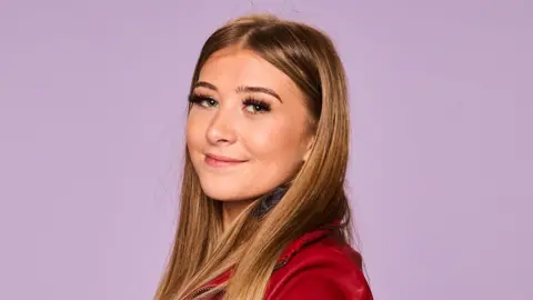 A woman with long brown hair poses for a photograph. She wears a red top and smiles at the camera.