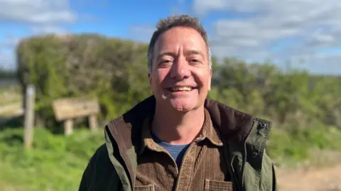 George Carden/BBC Patrick clarke wearing a green jacket and corduroy shirt smiling at the camera in the countryside