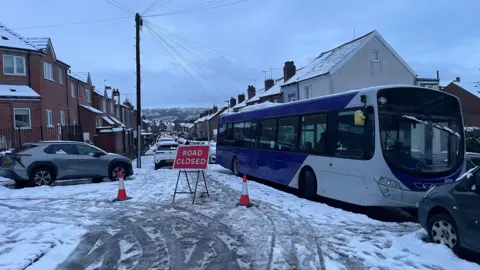 Peter Spencer/BBC A residential street covered in snow and ice, creating a slippery surface. In the foreground, there is a red sign that reads “ROAD CLOSED”, supported by a metal frame and flanked by two orange traffic cones. Behind the sign, a large blue and white bus is positioned diagonally across the street, appearing stuck or unable to move due to the icy conditions. Several parked cars line both sides of the street, and the houses are traditional brick homes with snow on their roofs.