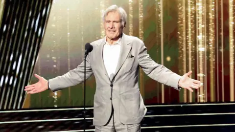 Reuters Harrison Ford es honrado en los Disney Legends Awards 2024 durante la Disney's D23 Expo en Anaheim. Harrison es un hombre de 82 años con cabello gris. Llevaba un traje gris claro sobre una camisa blanca y estaba en el escenario con los brazos extendidos.