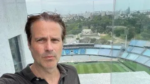 Chris Lee Chris Lee takes a selfie standing in a building that overlooks a football stadium. He has short dark hair that has been pushed back by sunglasses on his head. He wears a dark polo top. 