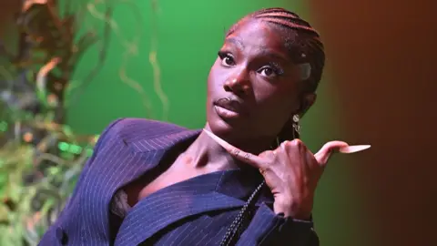 Getty Images Doechii, wearing a navy blue striped blazer, with her left hand making a "phone" sign. The background has a green light.