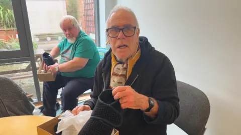 George King/BBC An elderly man wearing glasses and a black hoodie holding a pair of slippers while looking into the camera. 