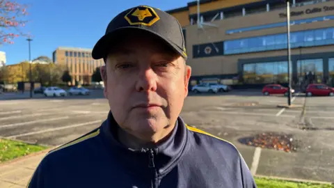 Dave Milton standing outside The Molineux Stadium. It is a huge building and we can see the Wolves logo. He is a middle aged white man. He is wearing a Wolves hat and a blue and golden jacket. 