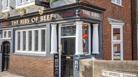 The entrance to the Ribs of Beef, which has a brick facade, white windows, a black door and a black banner with gold lettering advertising the pub's name.
