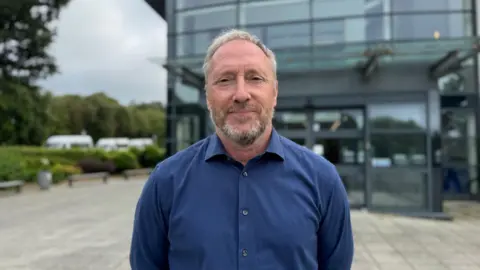 Paul Montague - a man with a short greying beard and short light grey hair. he is wearing a dark blue shirt and is looking at the camera smiling
