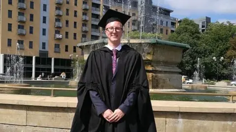 Matthew Brown is a young man wearing glasses standing in a gown and mortar board with his hands clasped in front of him. He is standing in front of a fountain 