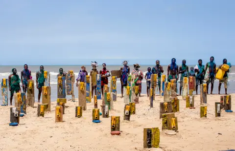 Nii Odzenma People standing near artwork using jerrycans by artist Serge Attukwei Clottey on a beach in Accra, Ghana