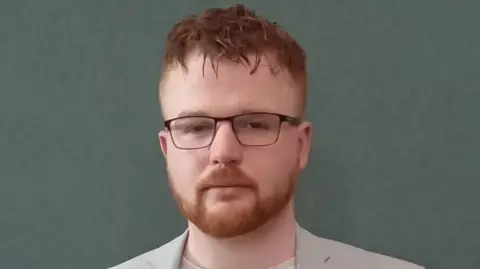 Head-and-shoulders image of Alex Clarke. He is looking into the camera. He has red hair and a beard. He is wearing black-rimmed glasses, a pale T-shirt and light grey blazer.