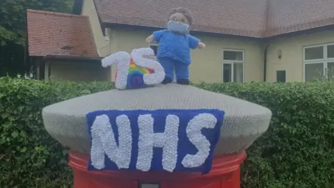 Lou Peden Knitted nurse in uniform wearing a mask stands next to a large "75" on top of a post box