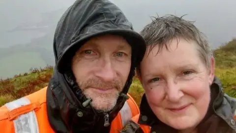 Sharon Rodhouse A man and woman smile at the camera in the rain